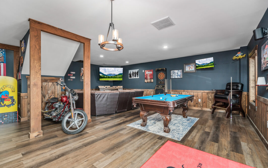 Photo of game room of home with pool table and large seating area for watching ballgames on the two mounted screens. The floor is wood and the wainscot is matching wood. The upper walls and ceiling are drywall.